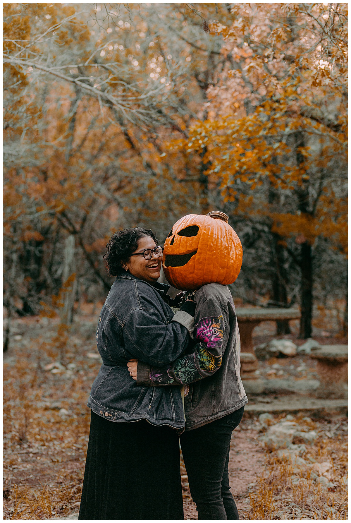 partners embrace in wooded area in the fall while one wears a pumpkin head by Austin portrait photographer