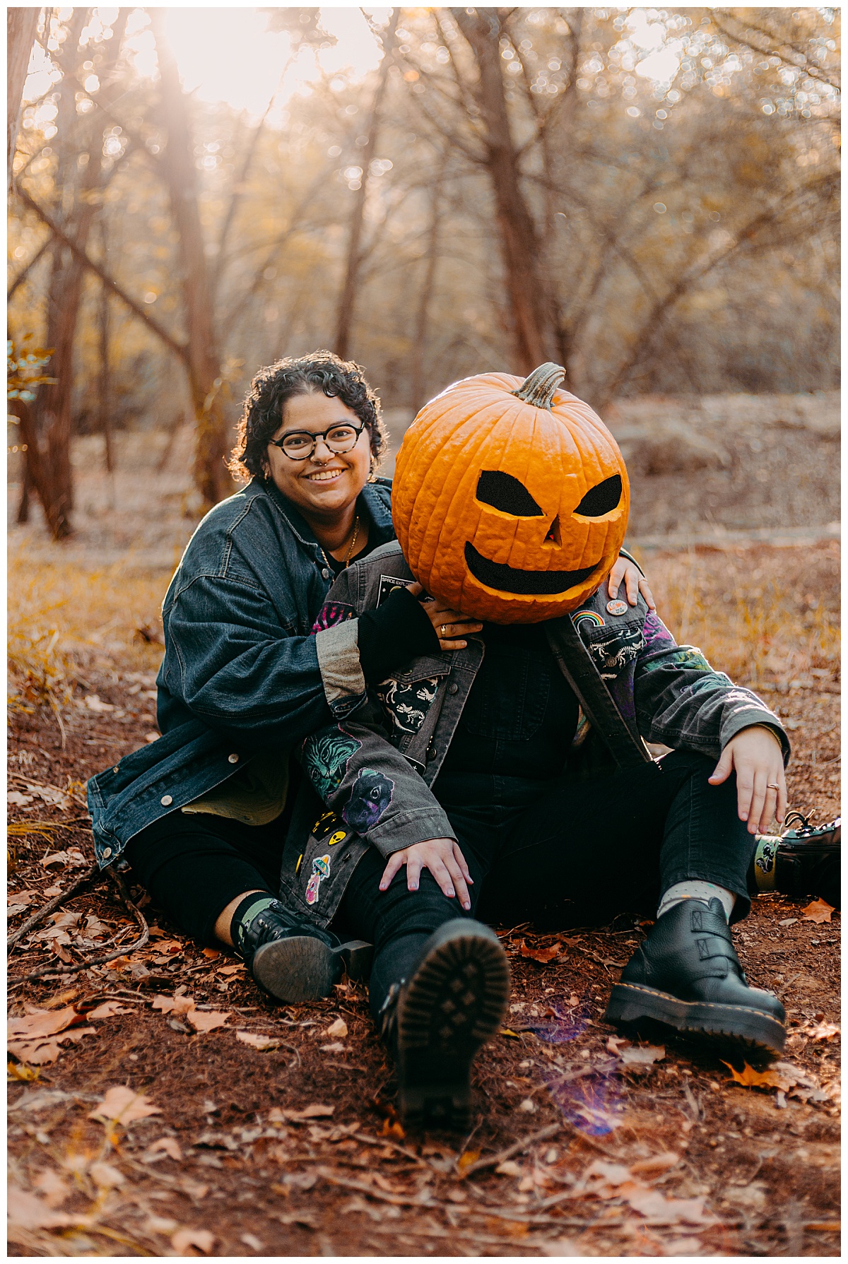 Wives sit on ground with fallen leaves wearing dark colors and a jack-o-lantern for Austin portrait photographer